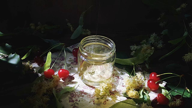 将蜂蜜倒入玻璃罐中，将一束菩提花和红樱桃放在木质表面。射线的阳光。黑暗的乡村风格。视频素材
