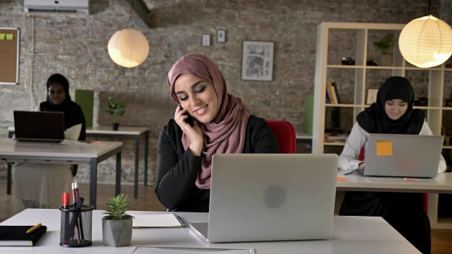 三个年轻的穆斯林妇女戴着头巾坐在现代化的办公室里工作，美丽的穆斯林妇女在打电话，面带微笑视频素材