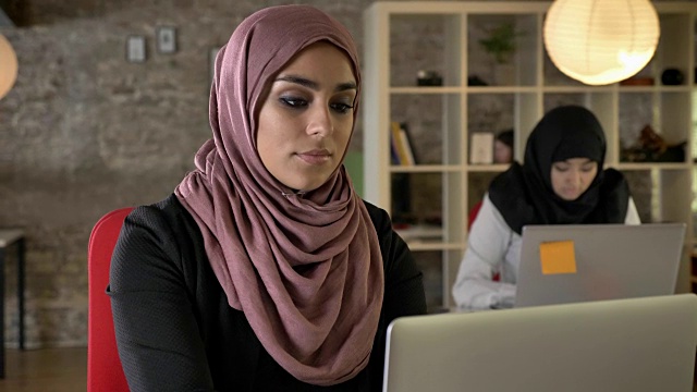 年轻的穆斯林妇女的肖像，严肃的工作，两个妇女戴着头巾，坐在笔记本电脑上打字在现代办公室，集中视频下载