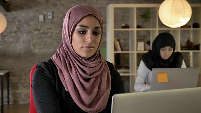 年轻的穆斯林妇女戴着头巾在工作，看着镜头，两个妇女坐在现代办公室的笔记本电脑上打字视频素材