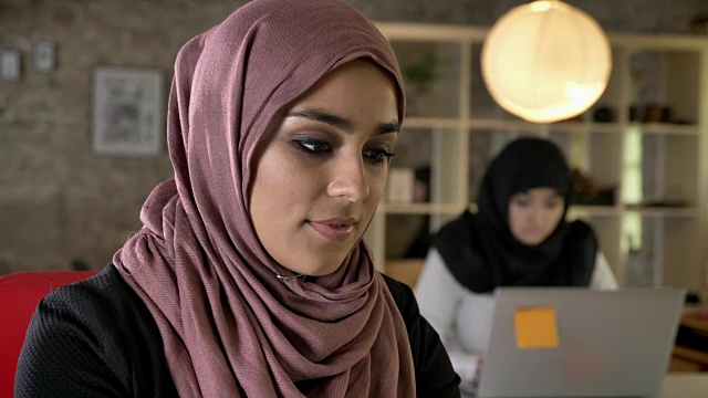 年轻的穆斯林妇女戴着头巾工作，面带微笑地看着镜头，两个妇女坐在现代办公室的笔记本电脑上打字视频素材