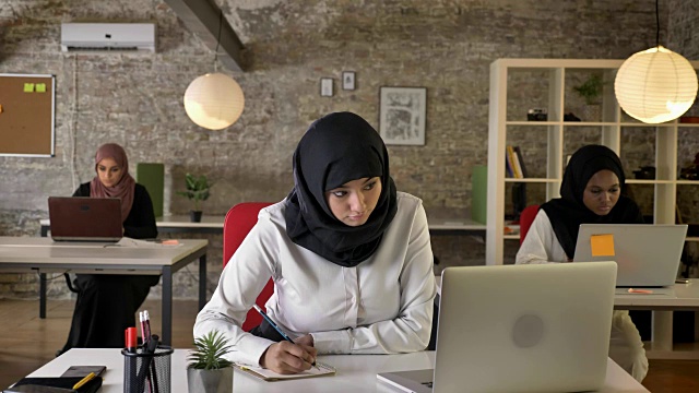 三个年轻的穆斯林妇女戴着头巾坐在现代办公室里工作，漂亮的女工人在纸上写字，忙碌视频素材
