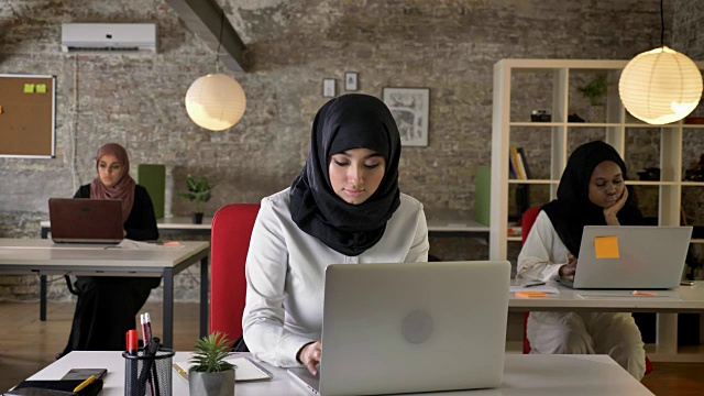 三个年轻的穆斯林妇女戴着头巾坐在现代化的办公室里工作，漂亮的女工人在笔记本电脑上打字，全神贯注视频素材