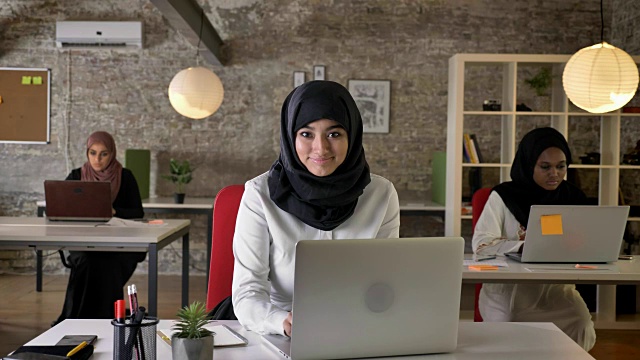 三个年轻的穆斯林妇女戴着头巾坐在现代办公室里工作，伊斯兰女工作者在笔记本电脑上打字，对着镜头微笑视频素材