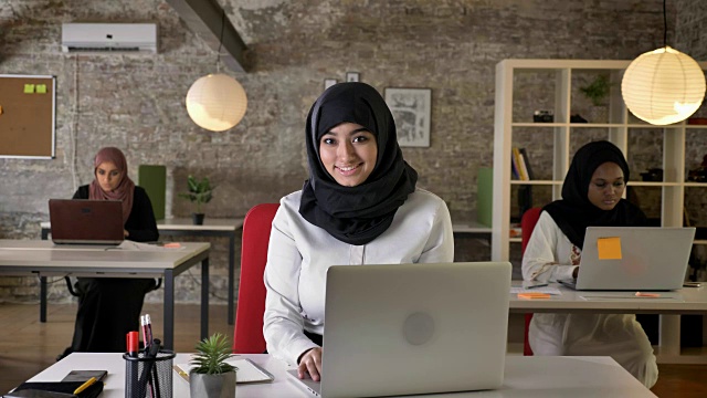 三个年轻的穆斯林妇女戴着头巾在现代办公室工作，漂亮的女性工作者在笔记本电脑上打字，对着相机微笑，开心视频素材