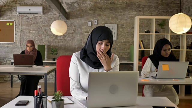 三个年轻的穆斯林妇女戴着头巾坐在现代办公室里用笔记本电脑工作，穆斯林妇女打着哈欠，疲惫不堪视频素材