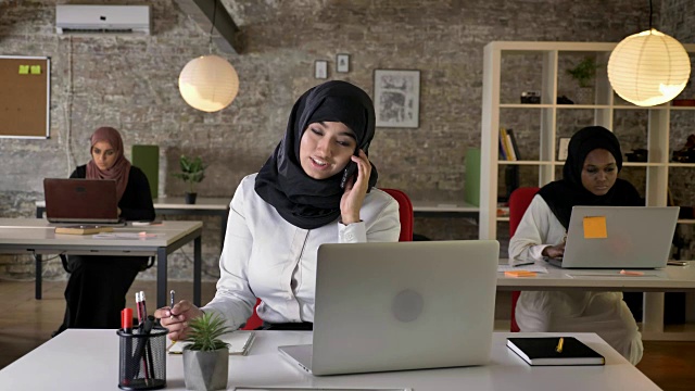 三个年轻的穆斯林妇女戴着头巾坐在现代办公室的笔记本电脑上打字，美丽的穆斯林妇女在打电话，忙碌视频素材
