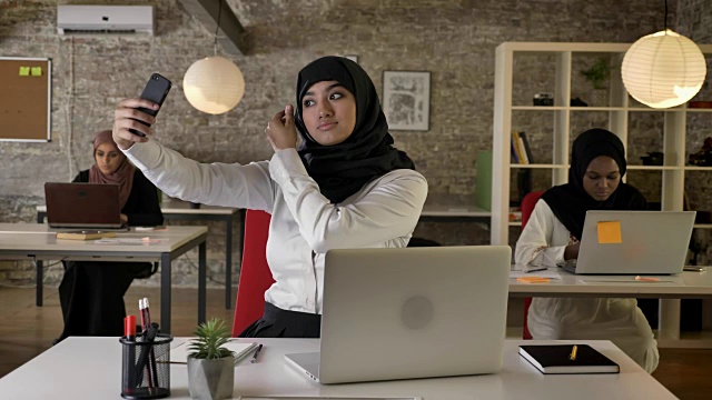 三名年轻的穆斯林妇女戴着头巾在现代办公室工作，而其他穆斯林妇女在用智能手机自拍视频素材