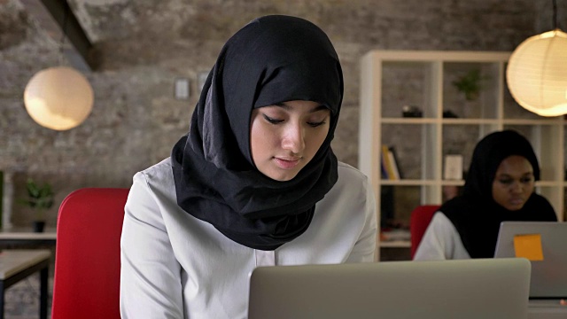 年轻的穆斯林妇女戴着头巾在工作，微笑着看着镜头，两名妇女坐在现代办公室里视频素材