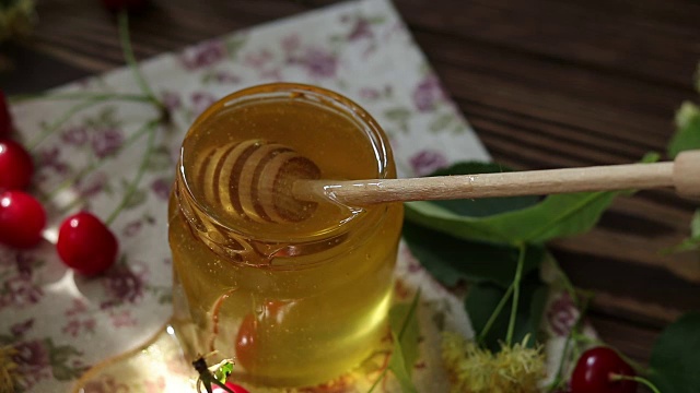 打开玻璃罐液体蜂蜜和蜂蜜勺，一束菩提花和红樱桃在木制表面。射线的阳光。黑暗的乡村风格。视频素材