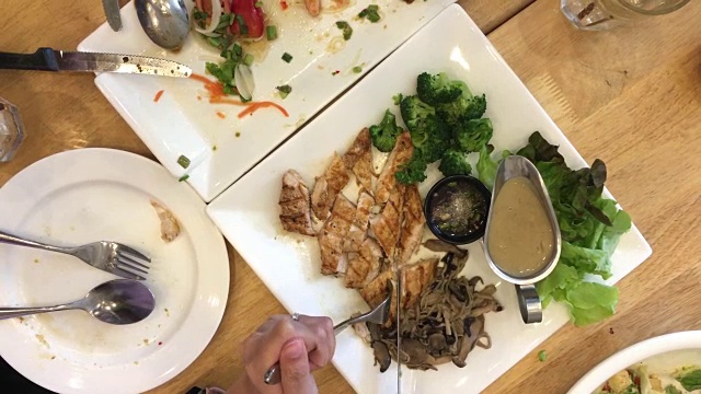 女人手拿刀叉切烤牛排在白色的盘子和食物，另一个是完成在餐馆的木桌上。前视图。视频片段。视频下载