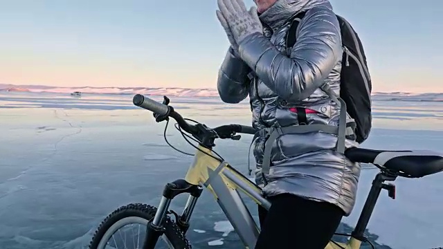 女人在冰上骑自行车。这个女孩穿着银色的羽绒服，背着自行车背包，戴着头盔。女人在用摩擦加热她的手。视频素材