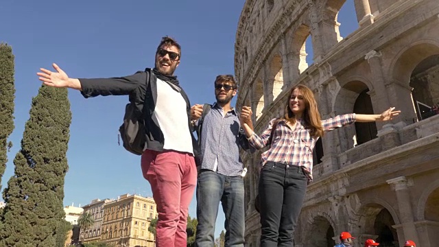三个年轻的朋友游客站在罗马圆形大剧场前的台座上，用背包、太阳镜、快乐、美丽的女孩、长发、慢镜头拍了一些滑稽搞笑的照片视频素材