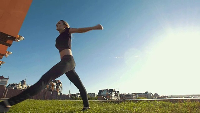 苗条的年轻女子表演杂技跳跃在草地上的背景城市景观视频素材