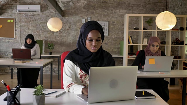 年轻的非洲穆斯林妇女戴着头巾在笔记本电脑上打字和咳嗽，三个美丽的穆斯林妇女在现代办公室工作视频素材