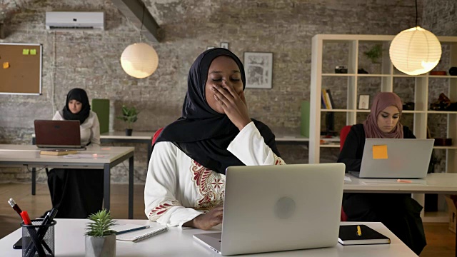 年轻的黑人穆斯林妇女戴着头巾在笔记本电脑上工作，打着哈欠，疲惫不堪，三个美丽的穆斯林妇女坐在现代化的办公室里视频素材