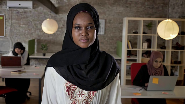 年轻的非洲穆斯林妇女戴着头巾站在镜头前，关切地看着三个在现代办公室里用笔记本电脑工作的妇女视频素材