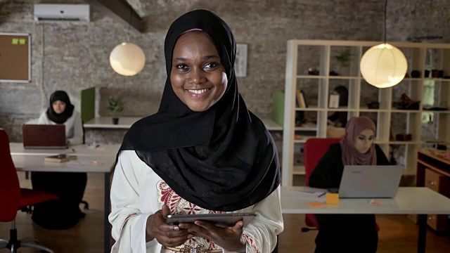 年轻的穆斯林黑人妇女戴着头巾使用平板电脑，看着相机，微笑着，伊斯兰商务妇女在现代办公室的笔记本电脑上打字视频素材