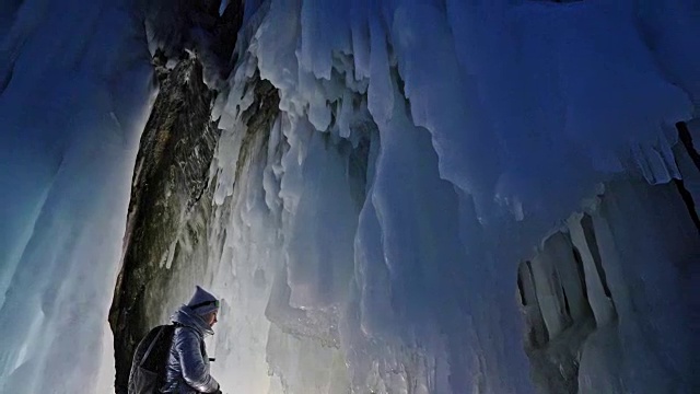 贝加尔湖冰洞上的女游客。冬岛之旅。女孩背包客正在冰洞中行走。游客看着美丽的冰洞。徒步旅行者穿银色夹克，背包，led灯。视频下载