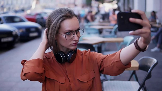 一个时髦的女孩在街中间用手机拍了张照片，整理她的头发，寻找一个角度。脖子上戴着耳机。模糊的背景上的汽车和人视频素材