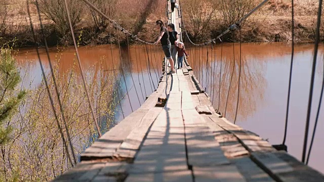 年轻的家庭妈妈、爸爸和女儿走在河上的老吊桥上。视频下载