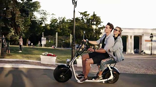 自信的男人在蓝色衬衫驾驶摩托车，而他的女朋友拥抱他。和朋友在公园边骑摩托车的神奇年轻女人视频下载