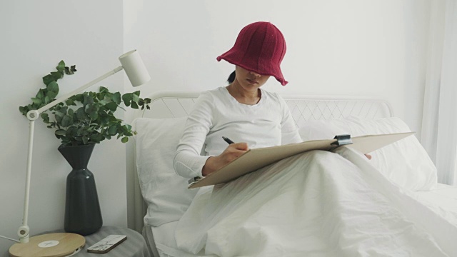 戴红帽子的女艺术家正在她的白色卧室里为她的休闲活动画画和素描视频下载