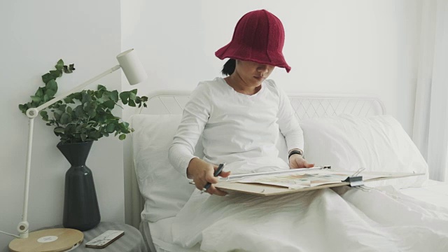戴着红帽子的女室内艺术家正在她的白色卧室里为她的休闲活动画画和素描视频下载