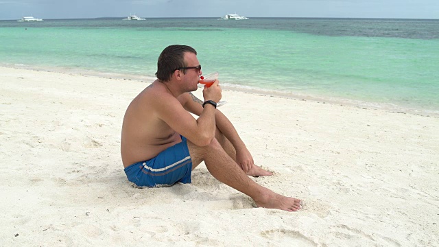 一个男人坐在海边的海滩上喝着鸡尾酒视频素材