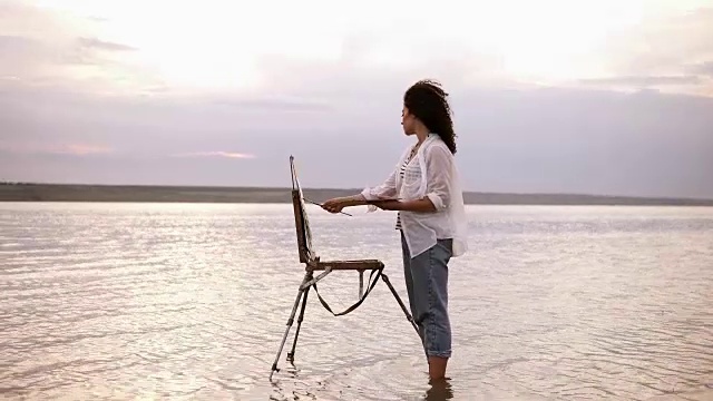 美丽的观点，一个年轻卷曲的黑发女子站在水与画架，并绘制她周围的风景。穿着蓝色的牛仔裤和白色的衬衫。完整的长度视频下载