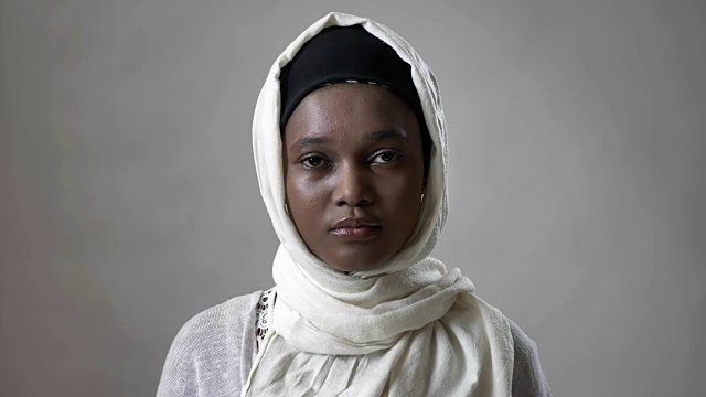 年轻的非洲穆斯林女孩戴着头巾看着镜头，宗教观念，灰色的背景视频下载