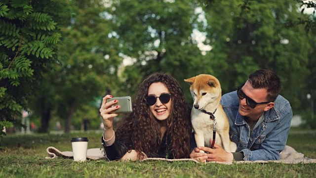 一名年轻女子和她的男友和宠物狗躺在公园的草地上自拍，可爱的动物在打喷嚏和舔鼻子，人们在笑。视频下载