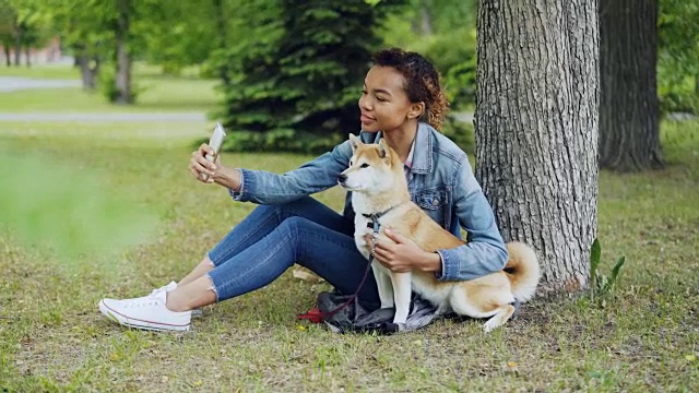 迷人的非洲裔美国女孩正在城市公园与可爱的狗狗自拍，拥抱和爱抚美丽的动物。现代科技、宠物和自然理念。视频素材