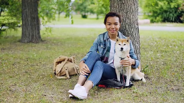 非裔美国女孩爱狗主人坐在公园草地上和她美丽的宠物，微笑着看着相机。爱护动物、爱护自然的理念。视频素材