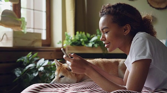 漂亮女生正在用智能手机触摸屏幕，和可爱的宠物狗躺在家里的床上大笑，动物享受着关爱。视频素材