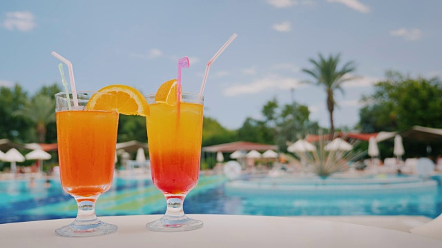 两杯清凉的鸡尾酒放在桌子上，背景是游泳池和棕榈树。天堂和豪华度假村视频下载