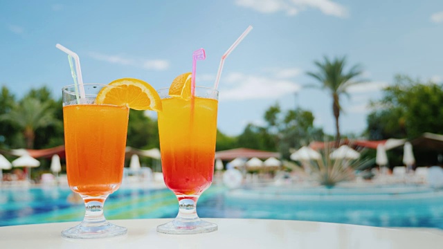 在游泳池附近的阳光躺椅上放松，用吸管喝两杯异国情调的鸡尾酒，蓝色的天空和绿色的棕榈树在泳池附近的背景视频素材