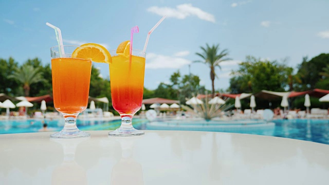 在游泳池附近的海滩度假，两杯彩色的酒精鸡尾酒在蓝天和异国情调的棕榈树视频素材