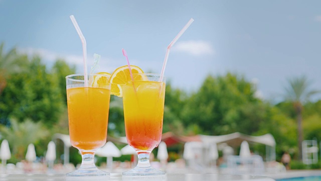 在蓝天的映衬下，泳池边的沙滩桌上放着两杯用吸管和橘片调制的橙色鸡尾酒视频素材