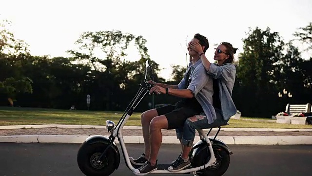 一对年轻的情侣正在约会，他们骑着一辆小型电子自行车在街上。穿休闲。金发女郎在骑马的时候闭上了他男朋友的眼睛。白天视频下载