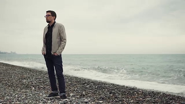 一个戴眼镜的男人站在海边多石的海滩上，看向别处视频下载