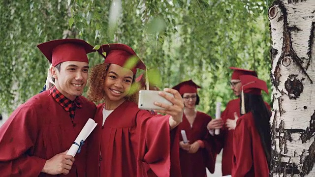 快乐的年轻人女孩和男孩在毕业典礼后穿着长袍和学士帽拿着学位证书自拍。照片和教育理念。视频素材