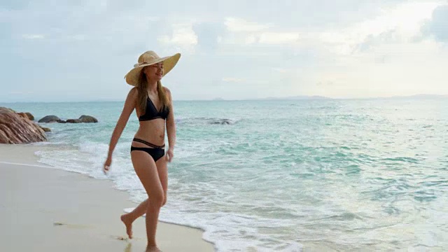 早上，一个女人戴着海帽，穿着黑色比基尼走在海滩上视频素材