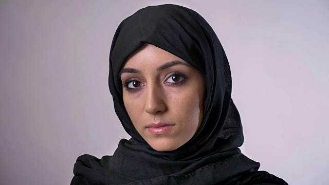 年轻严肃的穆斯林女孩戴着头巾看着镜头，灰色的背景视频下载