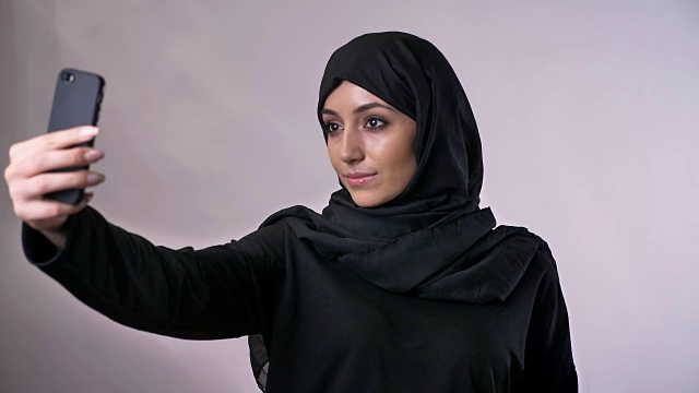 年轻甜美的穆斯林女孩戴着头巾在她的智能手机上自拍，手表照片，交流概念，宗教概念，灰色背景视频下载