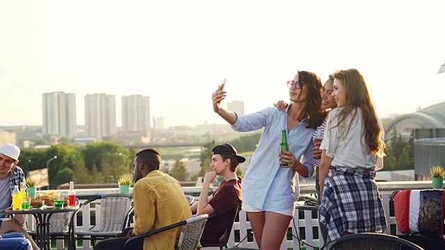 快乐的女性朋友们用智能手机自拍，大笑着站在屋顶上喝着瓶子里的饮料享受派对。科技、饮料、青年理念。视频素材