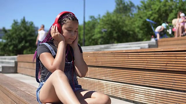 女孩戴着耳机坐在公园的长椅上听音乐。4 k视频下载