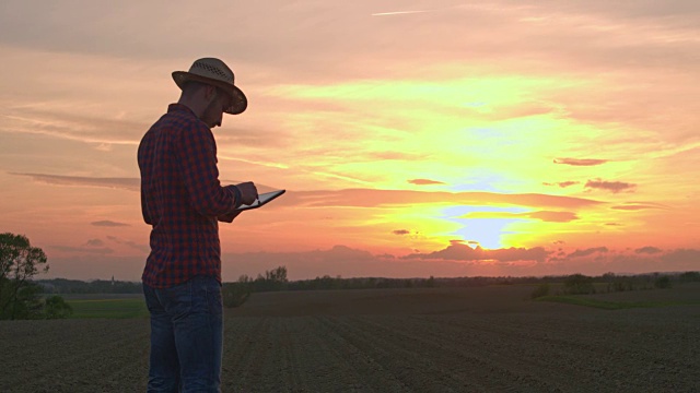 日落时分，法默在田园诗般的乡村耕地上使用数字平板电脑视频素材