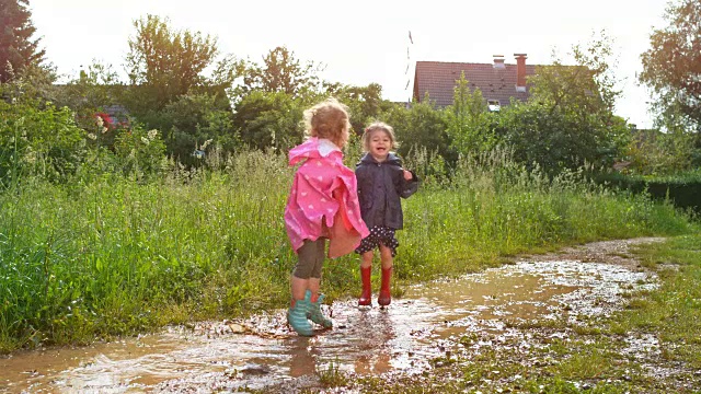 LD两个小女孩穿着雨靴和雨衣在泥泞的水坑里跳上跳下视频下载