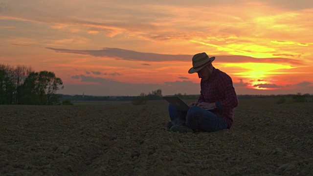 日落时分，法默坐在田园诗般的乡村耕地上使用笔记本电脑视频素材